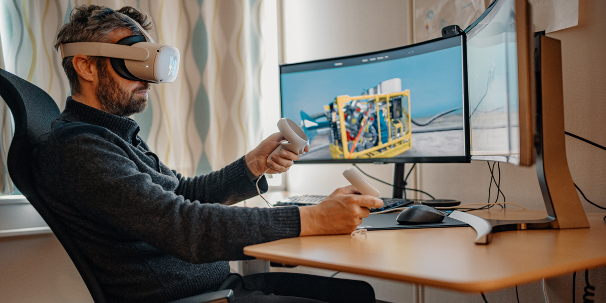 Engineer using virtual reality headset and controllers to simulate offshore drilling operations at a workstation with a wide curved monitor showing subsea equipment.